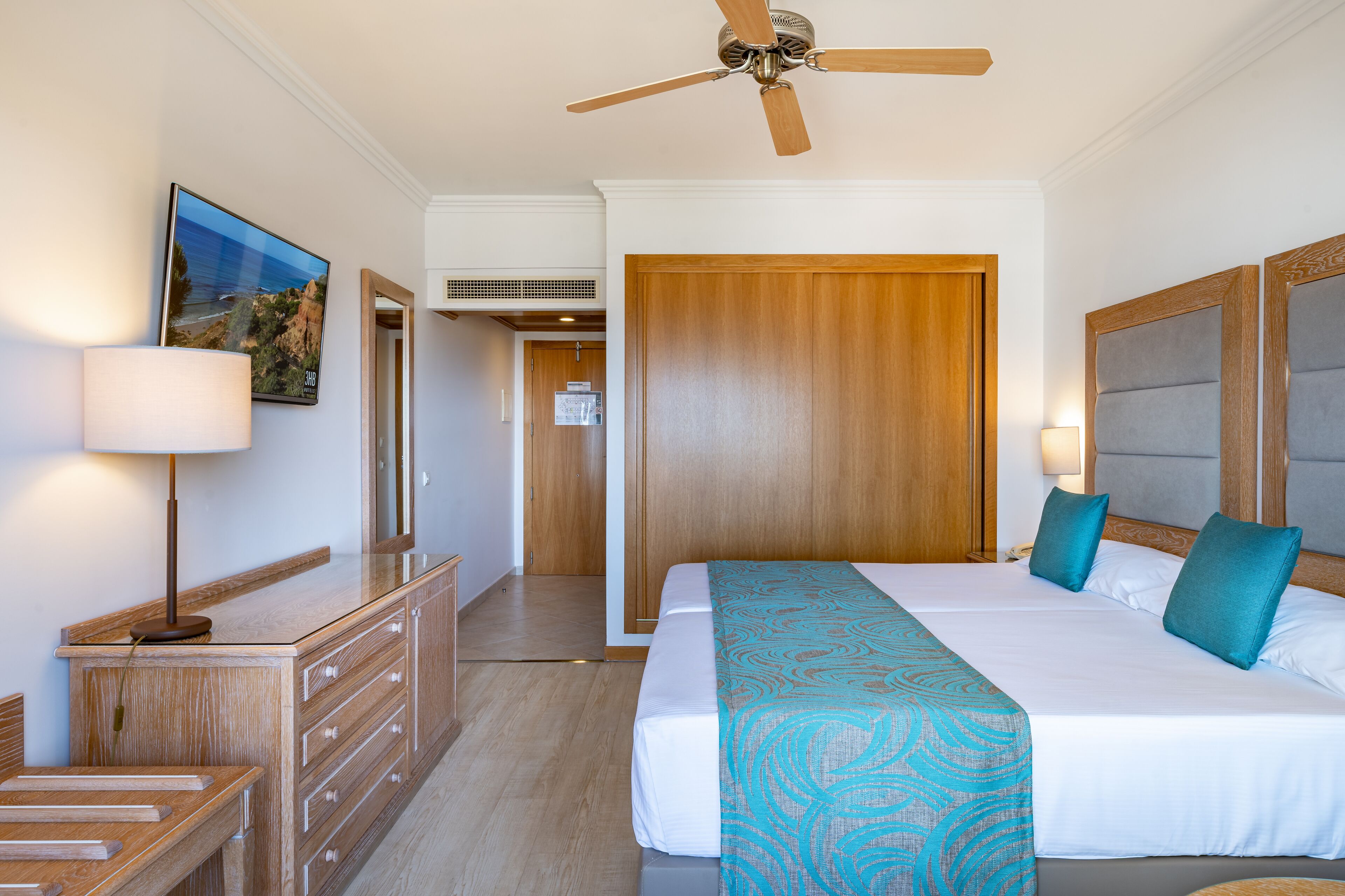 A hotel room with a bed, a desk, a chair, a ceiling fan, and a painting on the wall.