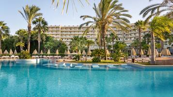 Una gran piscina con sillones de playa y sombrillas, rodeada de palmeras y un edificio de hotel al fondo.