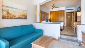 A modern hotel room with a teal sofa, a small wooden table, a painting of a beach, a kitchenette, and a wooden door leading to the bathroom. - 3HB Guarana (Albufeira)