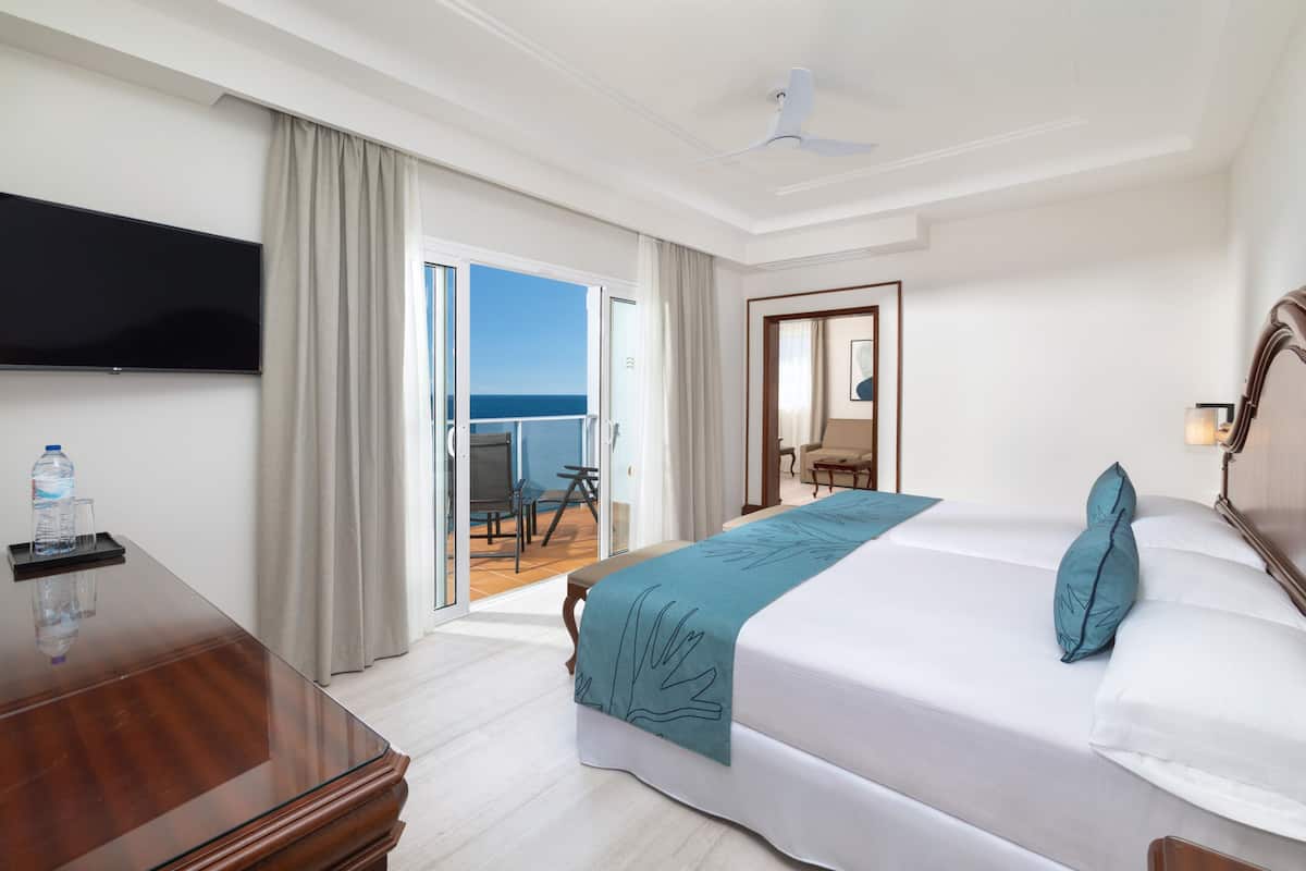 A hotel room with a bed, a desk, a TV, a balcony with a view of the sea, and a ceiling fan.