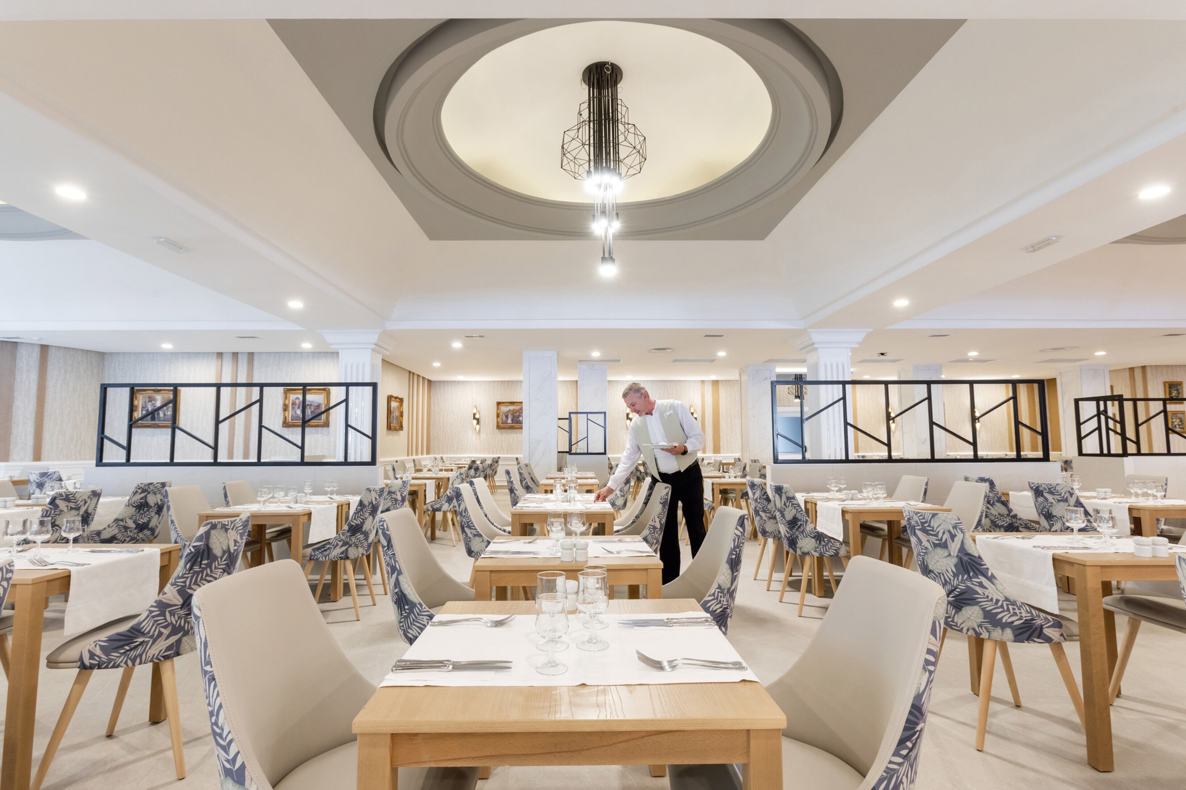 A spacious dining area with multiple tables and chairs, a waiter attending to guests, and a modern chandelier.