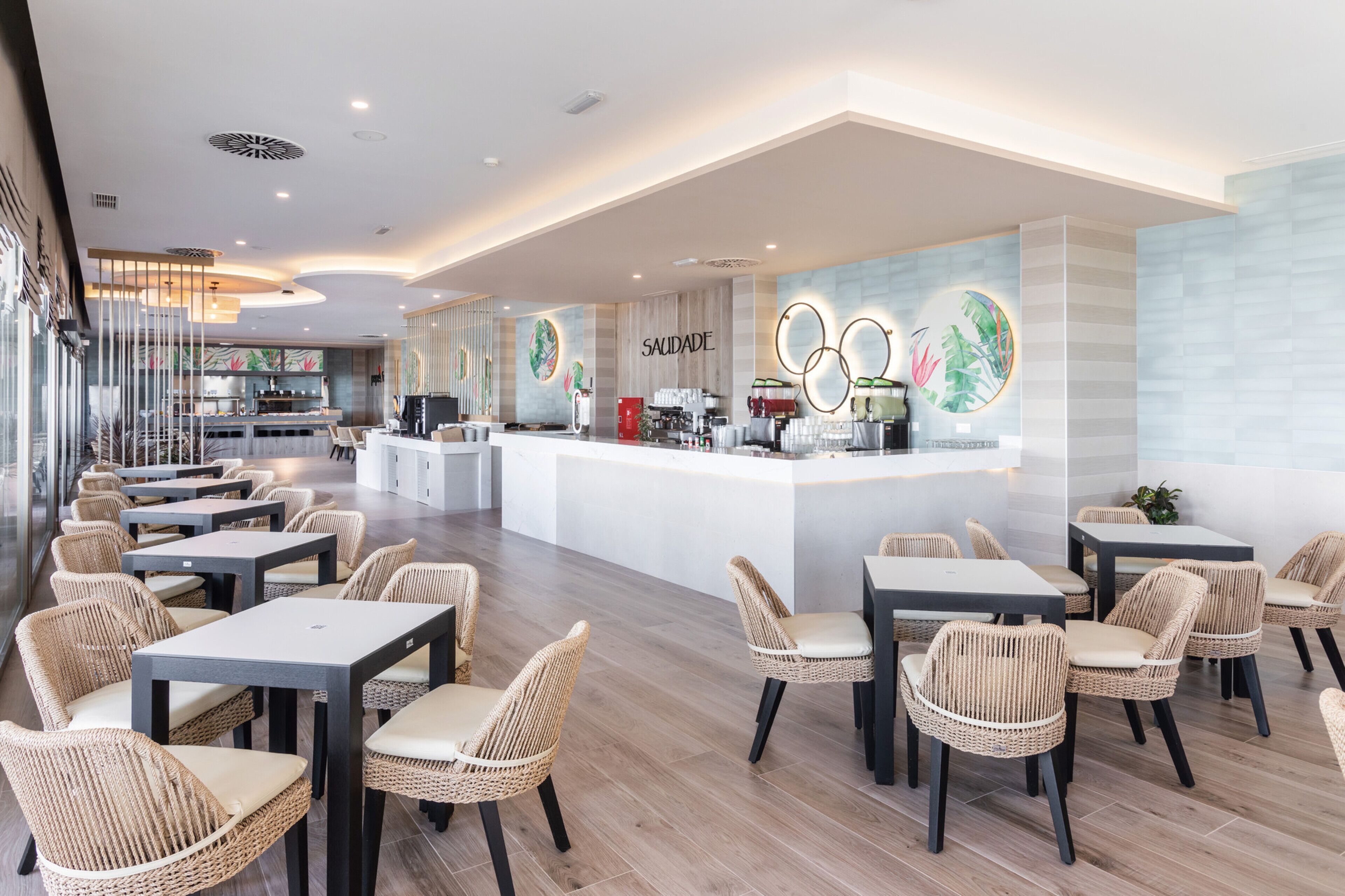 A modern cafe with wicker furniture, a central counter, and a mural with the word "SAUDADE".