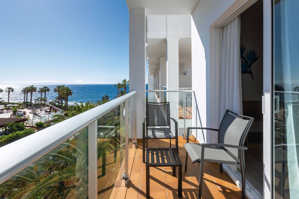 A balcony with a view of the ocean, a pool area, and palm trees.