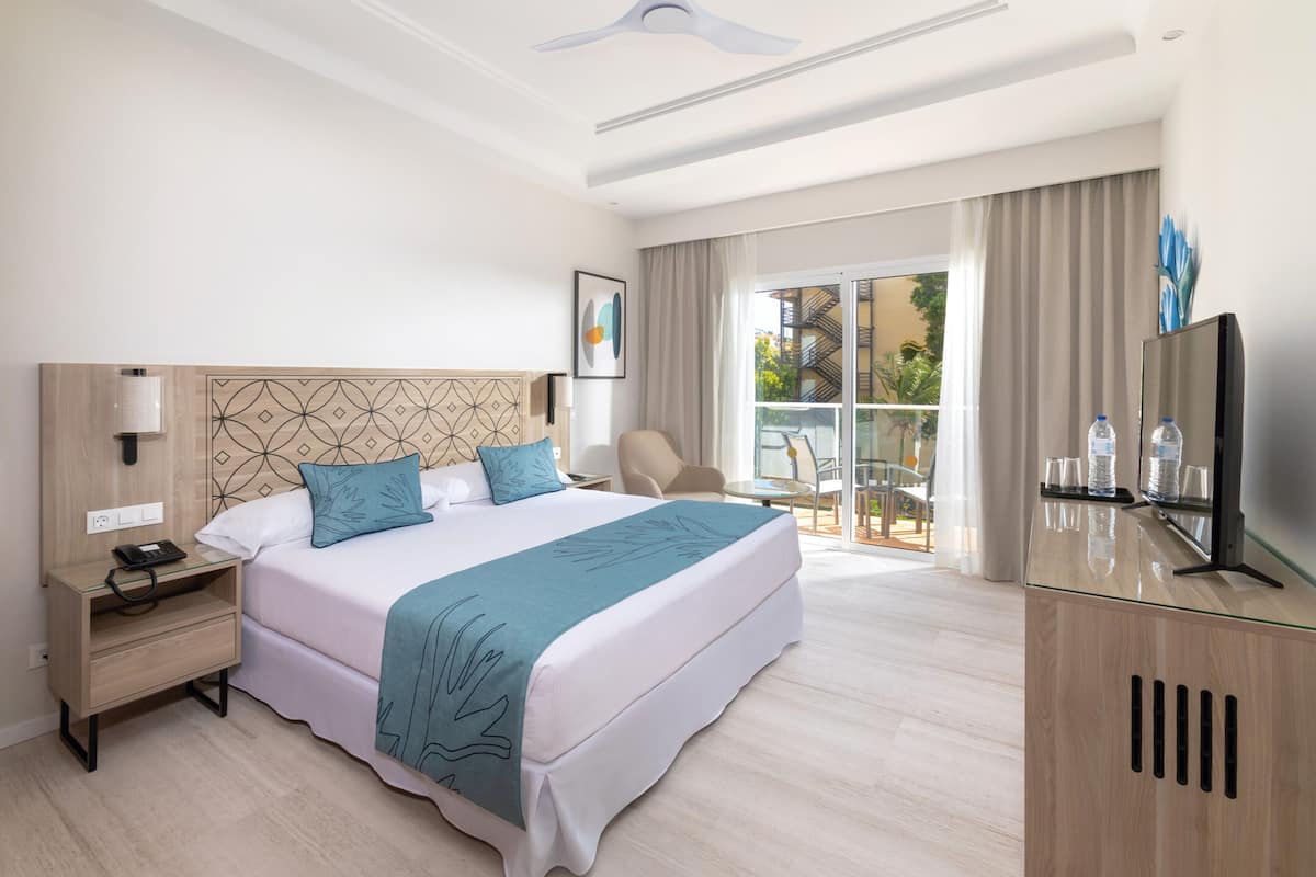 A hotel room with a large bed, a desk, a chair, a TV, and a balcony with a view of the city.