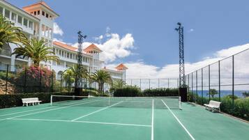 Een tennisbaan met een net, omringd door een hek, banken en palmbomen.