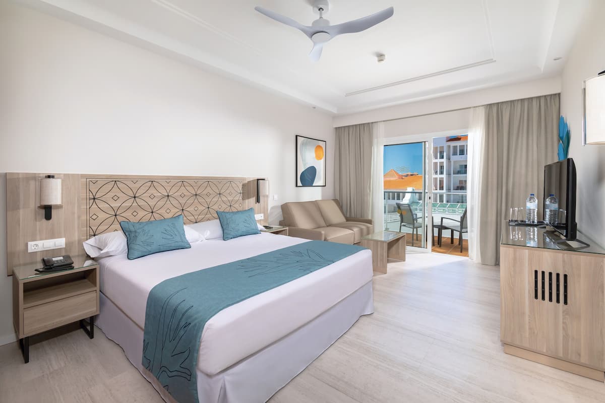 A hotel room with a large bed, a desk, a chair, a television, and a balcony with a view.