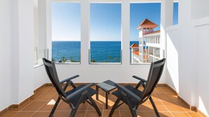A balcony with two chairs and a table, overlooking a clear blue sea and a building with a red roof. - Hotel Riu Madeira - All Inclusive (Santa Cruz)