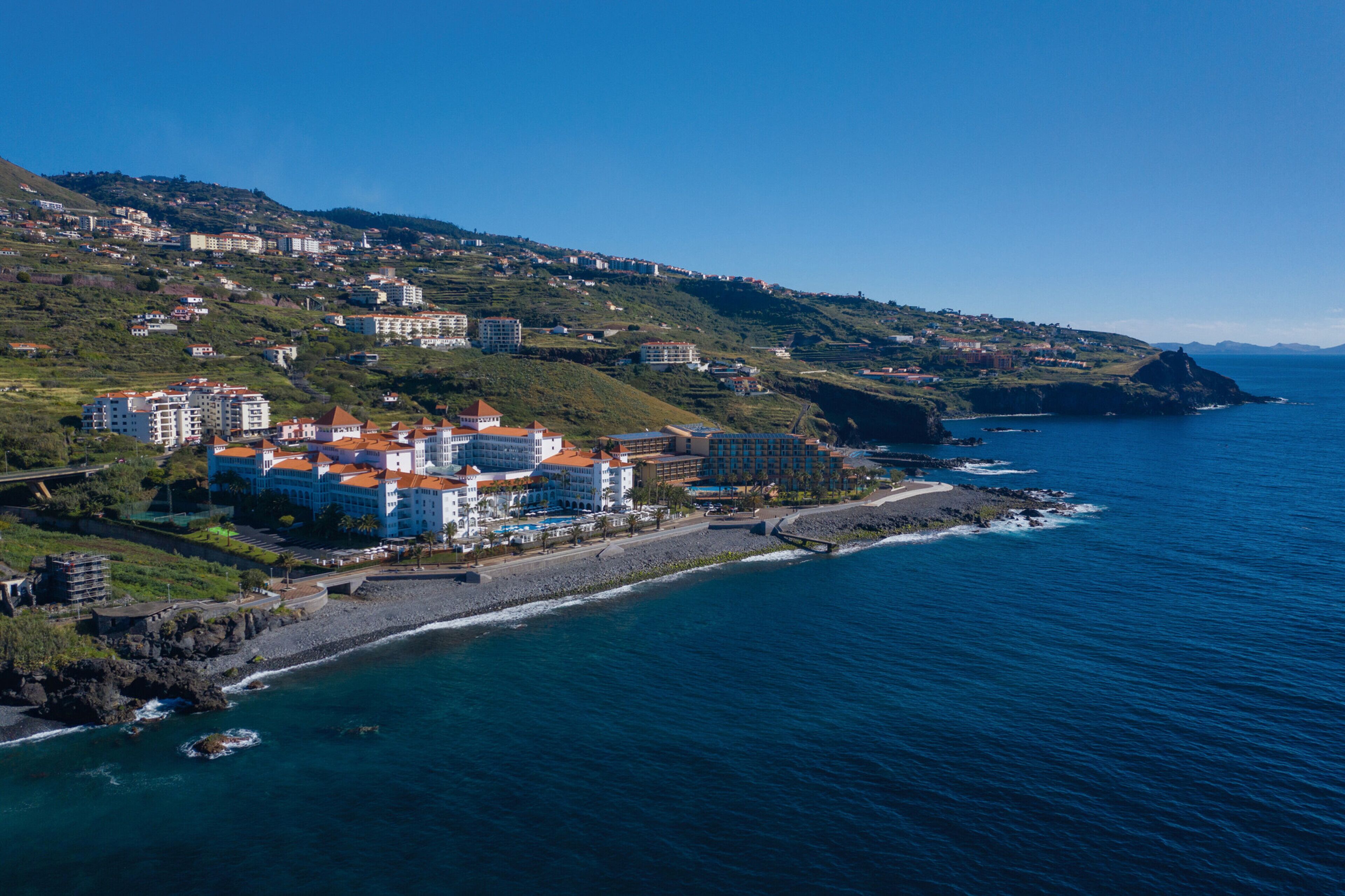 An aerial view of a coastal town with buildings along the shoreline, a beach, and a rocky outcrop in the distance.