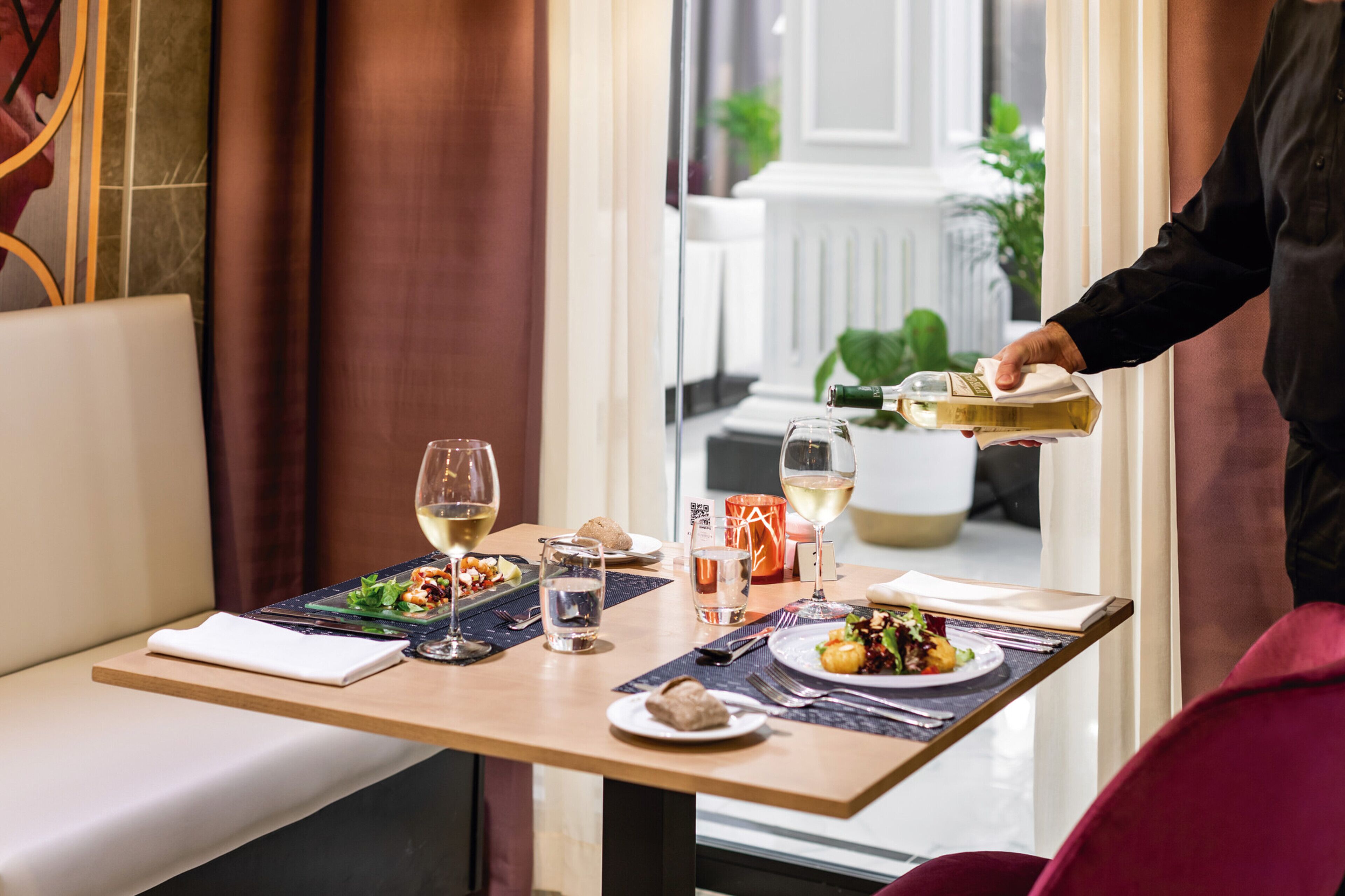 A dining table set with plates, glasses, and a bottle of wine, with a person serving food.