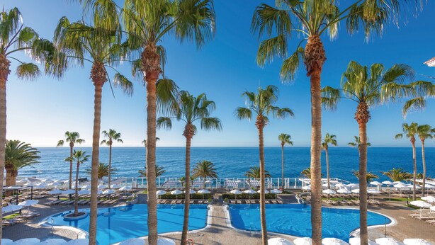 A pool area with multiple swimming pools, palm trees, and lounge chairs. - Hotel Riu Madeira - All Inclusive (Santa Cruz)