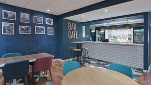 A bar café with a wooden table and chairs, a counter with a menu, and framed pictures on the wall. - Travelodge London Ealing (London)