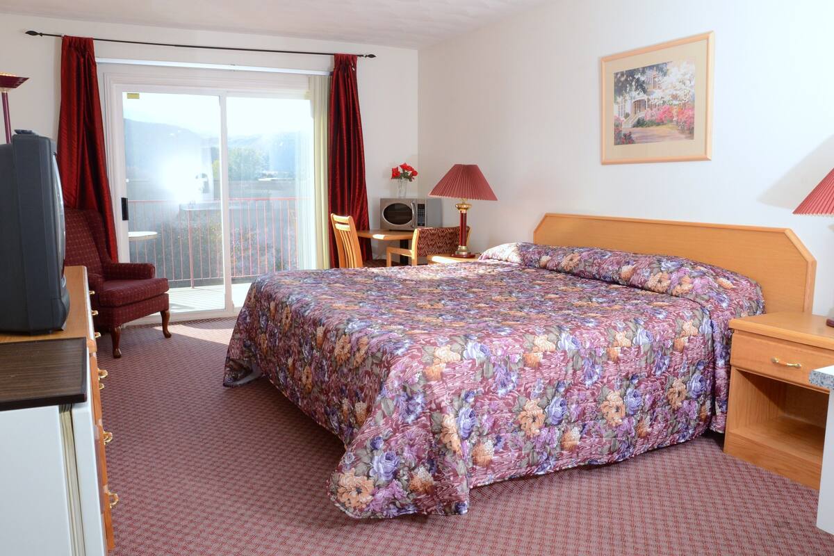 A hotel room with a bed, a desk, a chair, a TV, and a balcony with red curtains.