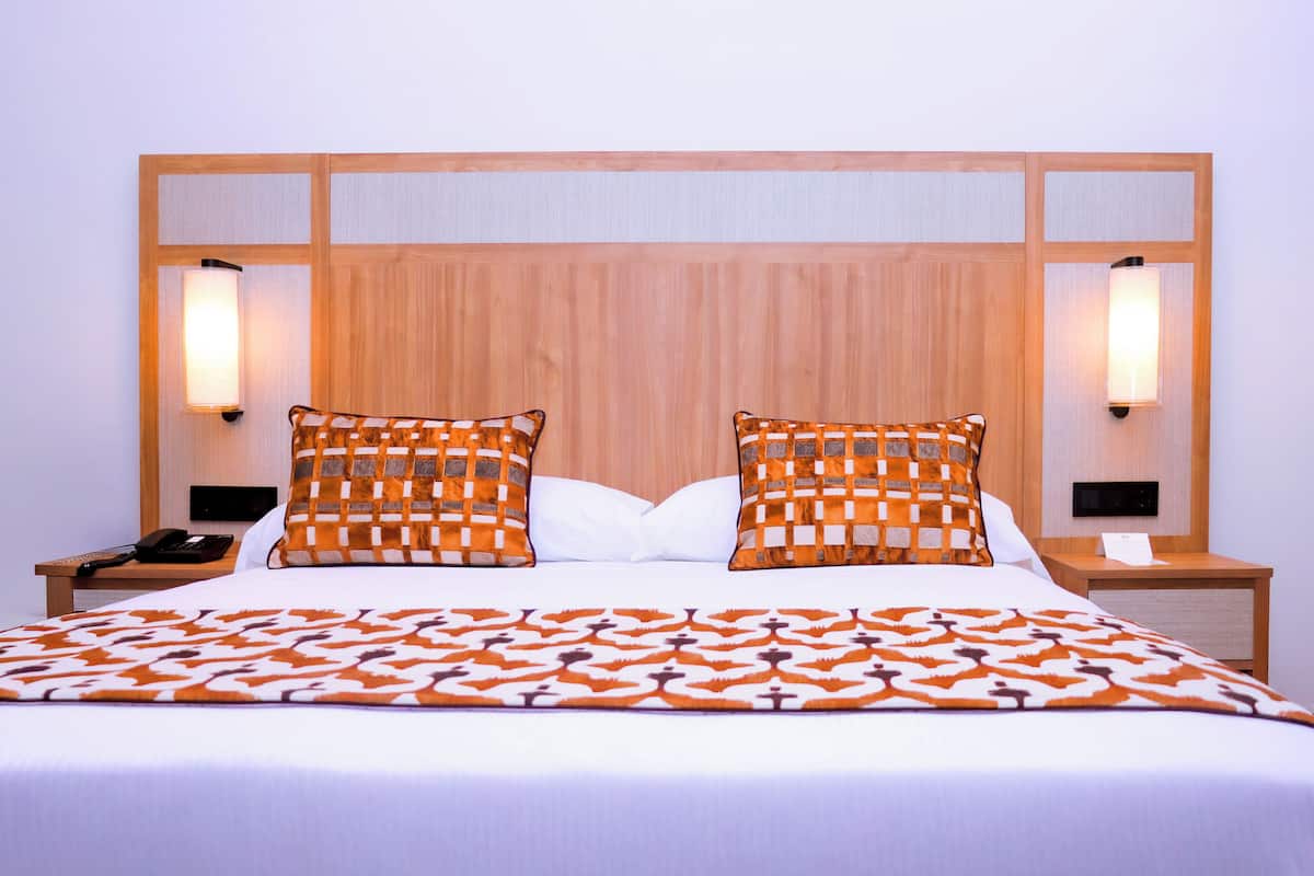 Hotellihuone, jossa on puinen sängynpääty, kaksi yöpöytävalaisinta, puhelin ja kuviollinen päiväpeite.