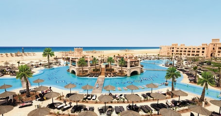 A resort with a central pool, palm trees, and beach umbrellas.