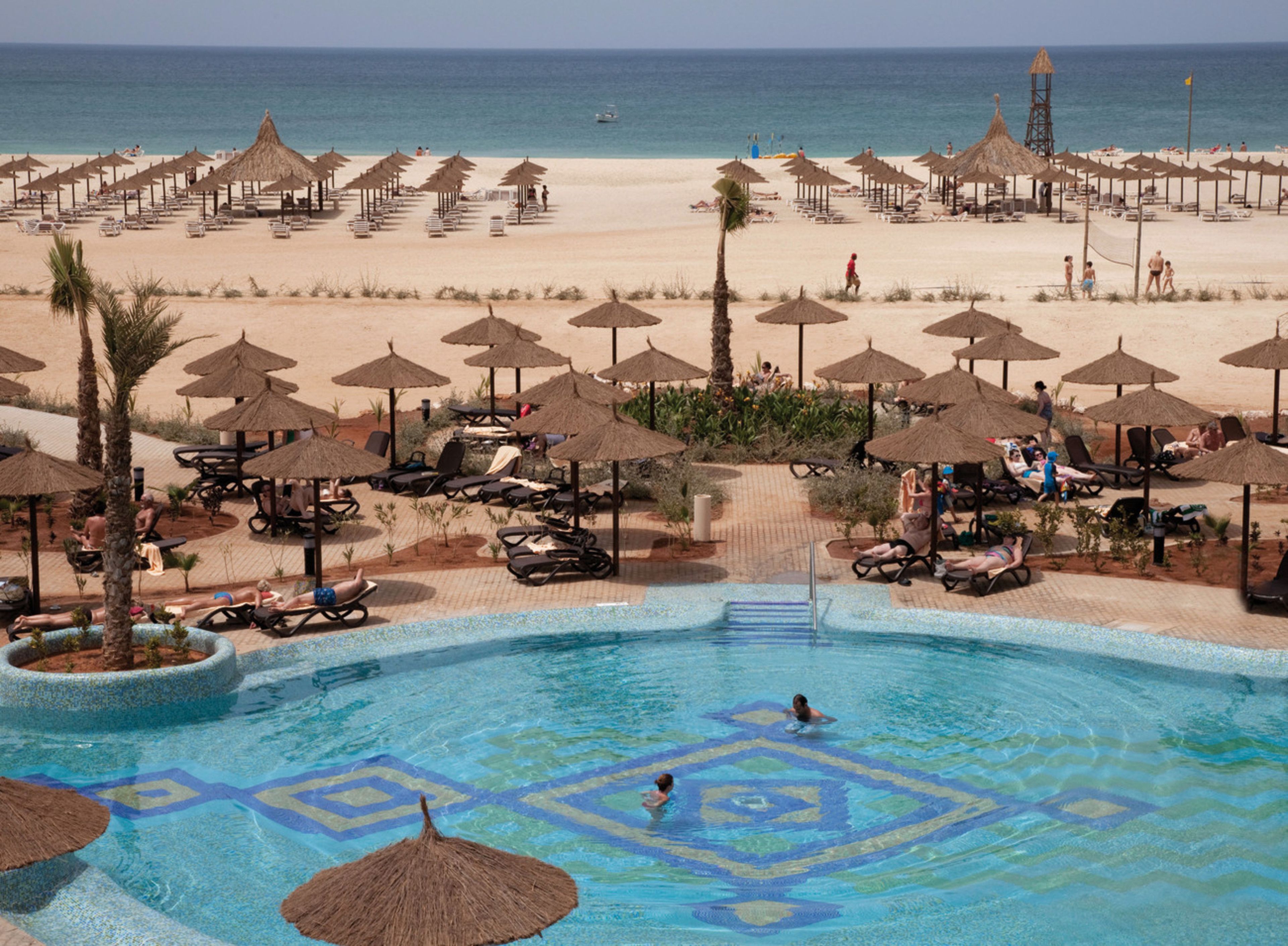 Un resort con piscina, sillas de descanso, sombrillas y una playa al fondo.