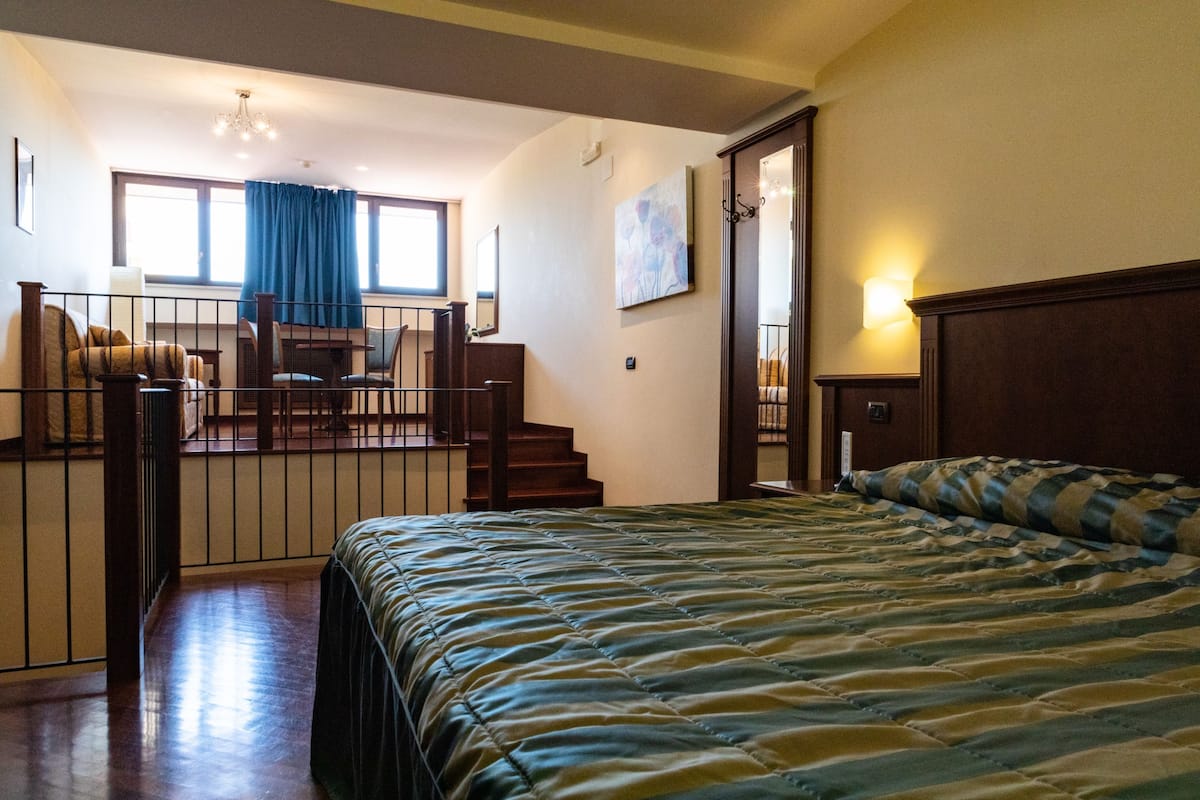 A bedroom with a large bed, a desk, and a chair. There is a staircase leading to an upper level.