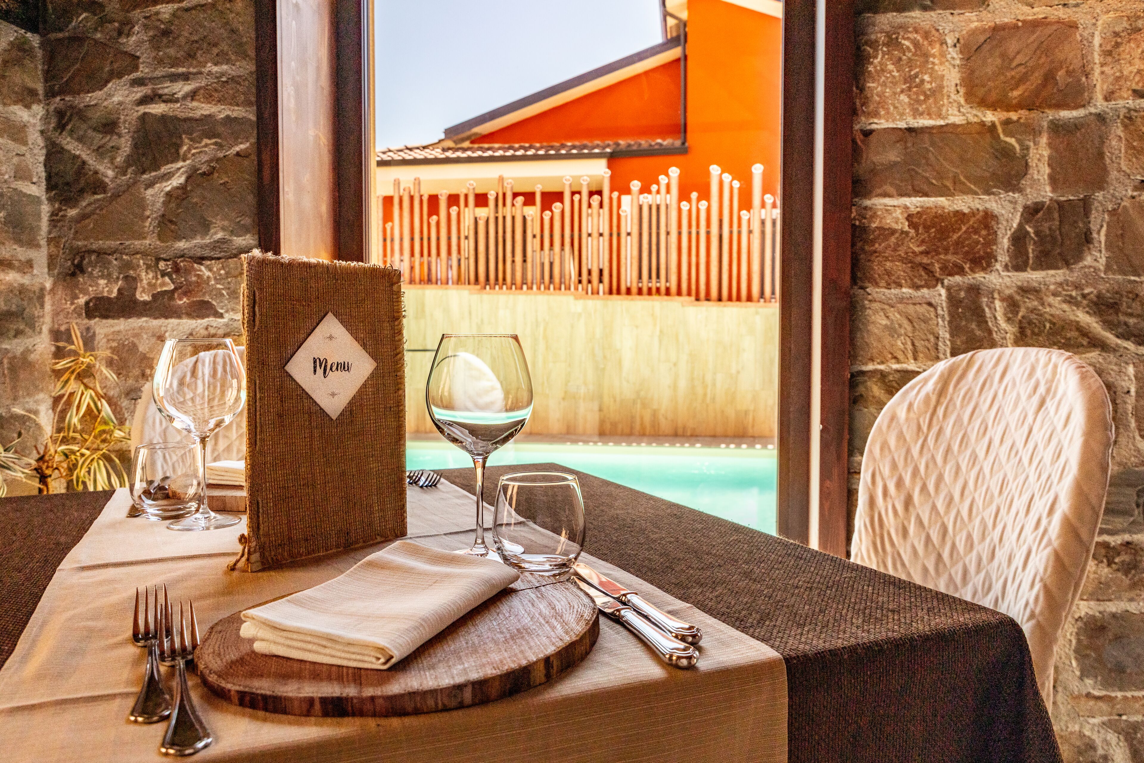 A dining table set with a menu, wine glasses, and cutlery, overlooking a pool with a wooden fence.