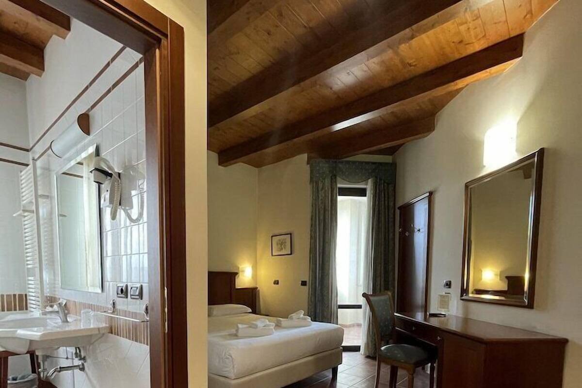 A hotel room with a wooden ceiling, a bed, a desk, a chair, a mirror, and a bathroom with a sink and shower.