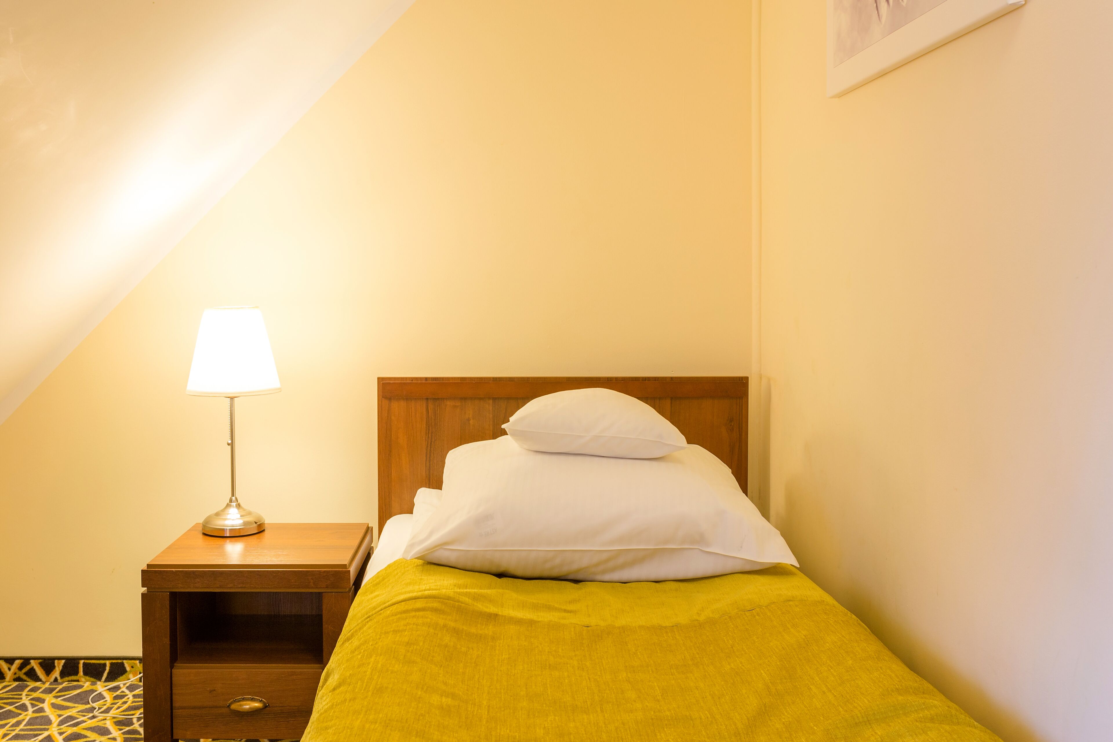 Un lit simple avec une tête de lit en bois, un oreiller blanc et une couverture verte. Il y a une table de chevet avec une lampe et un tiroir.