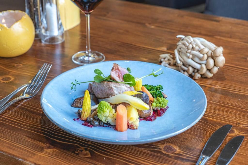Un plato de comida con diversos vegetales y un trozo de carne, acompañado de una copa de vino tinto y un salero sobre una mesa de madera.