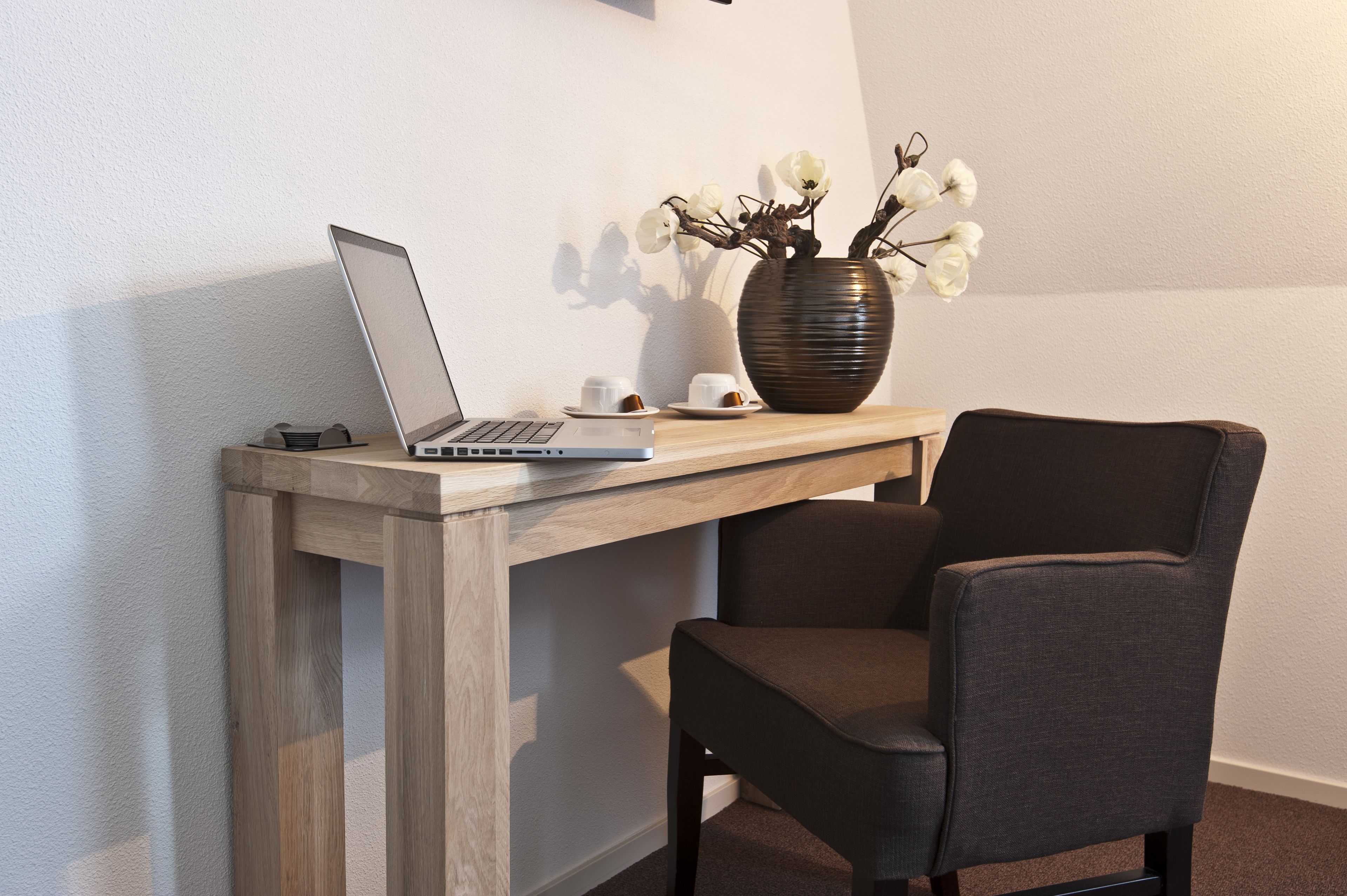 Ein Laptop steht auf einem Holztisch, daneben eine Vase mit weißen Blumen und zwei Tassen.