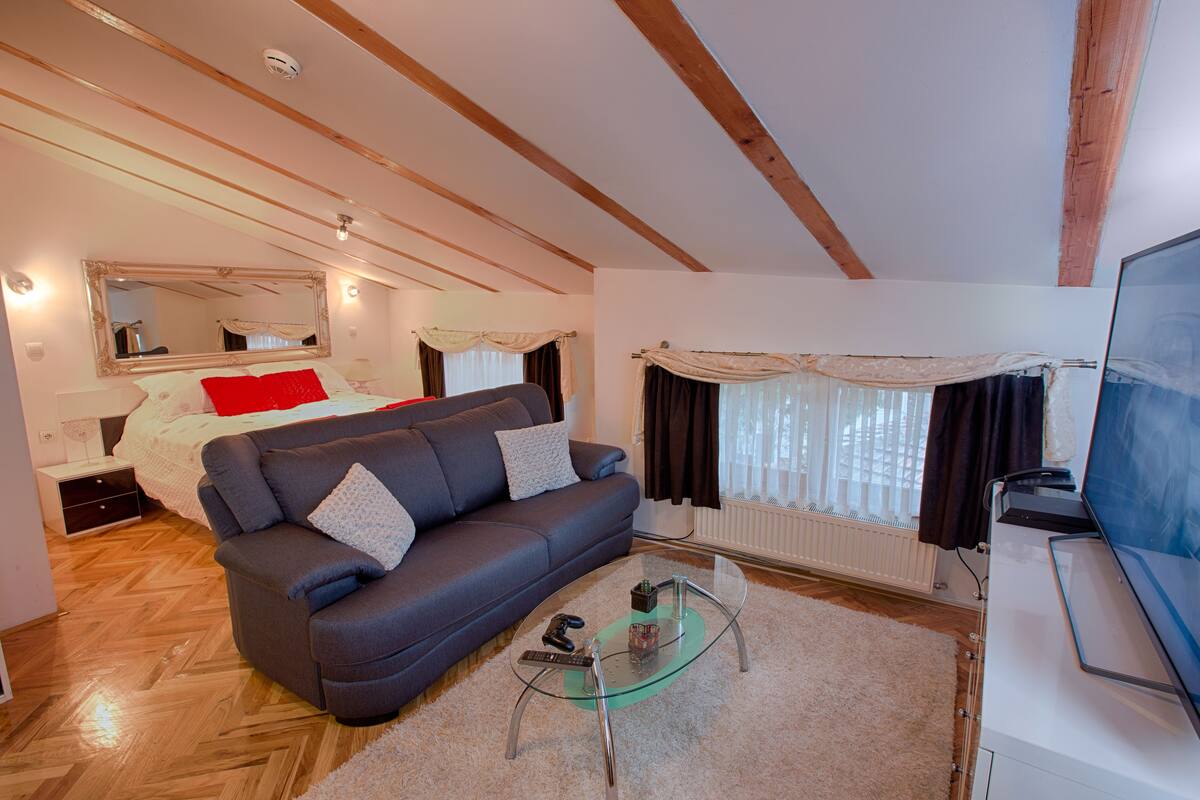 A modern living room with a sofa, a glass coffee table, a bed with a red pillow, and a large mirror.