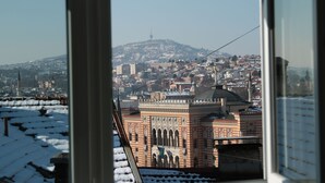 A view of a cityscape with a prominent building featuring arched windows and a flag, viewed through a window with snow on the ledge. - Hotel Herc (Sarajevo)