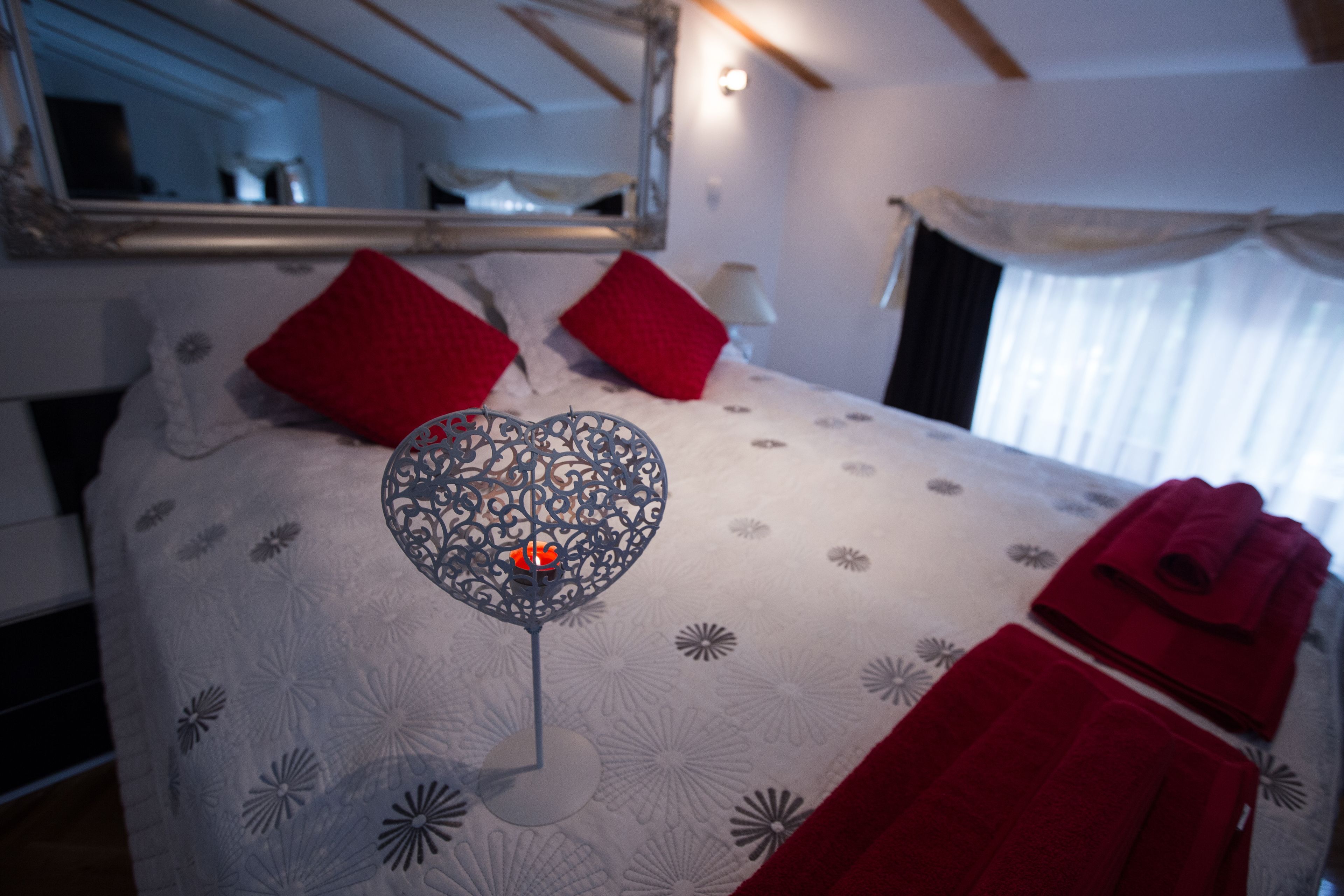 A bedroom with a bed, red pillows, a heart-shaped wall decoration, and a red towel.