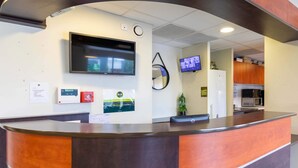 A reception area with a curved wooden counter, a mounted TV, a fire alarm, and a refrigerator in the background. - B&B HOTEL Agen Castelculier (Castelculier)