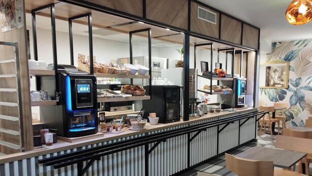 A modern cafe with a long counter, coffee machines, and seating area. - B&B HOTEL Agen Castelculier (Castelculier)