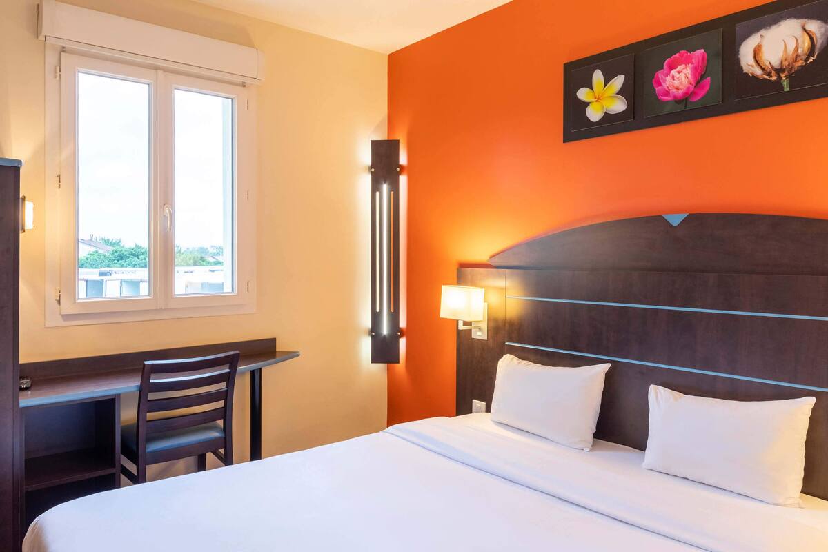A hotel room with a bed, a desk, and a chair. There is a window with blinds, a wall-mounted light, and a painting of flowers.