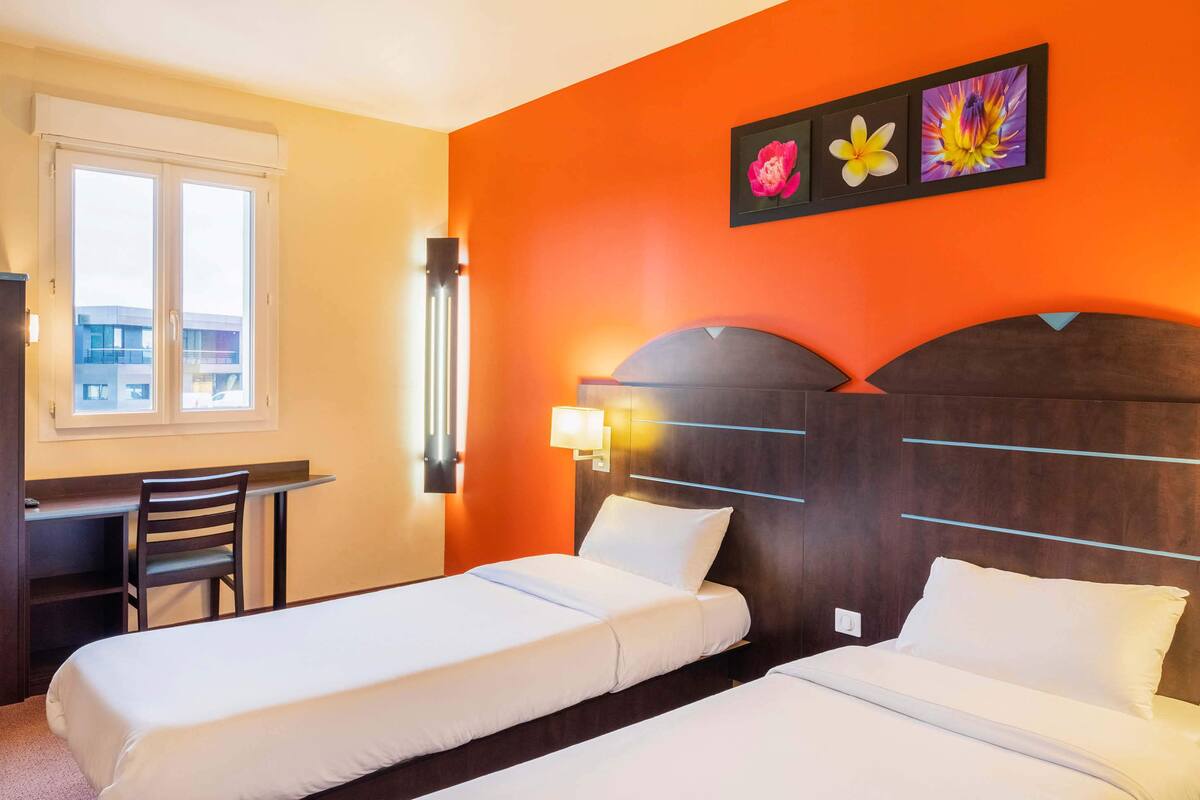 A hotel room with two single beds, a desk, and a chair. There is a window with a view of buildings outside.
