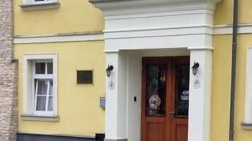 A building with a yellow facade, a central entrance with a wooden door, and two birds on a cobblestone ground.