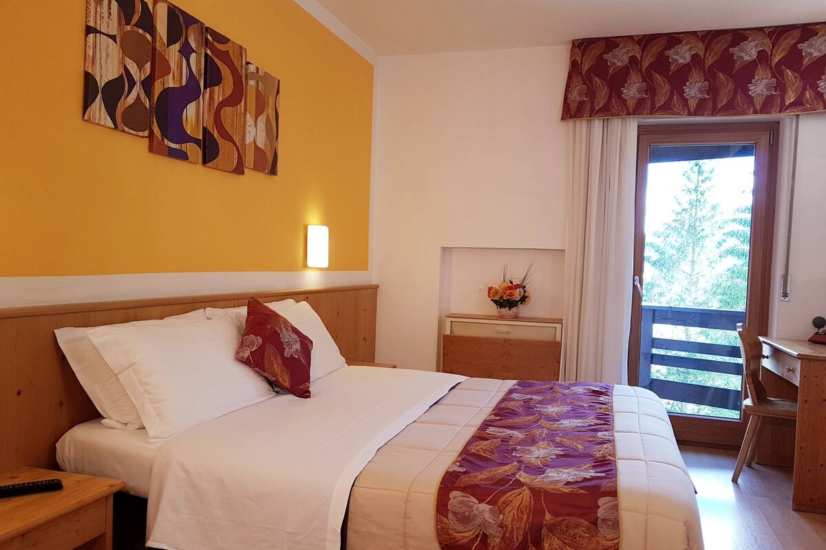 A hotel room with a bed, a nightstand, a desk, a window with a view of trees, and a wall with abstract art.