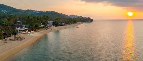 A beach with a row of houses along the shore, a calm sea, and a sunset in the background.