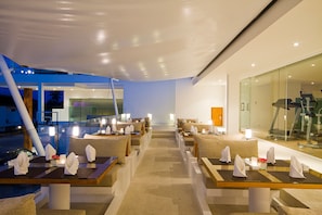 A modern outdoor dining area with multiple tables and chairs, a glass wall on one side, and a view of the street in the background.