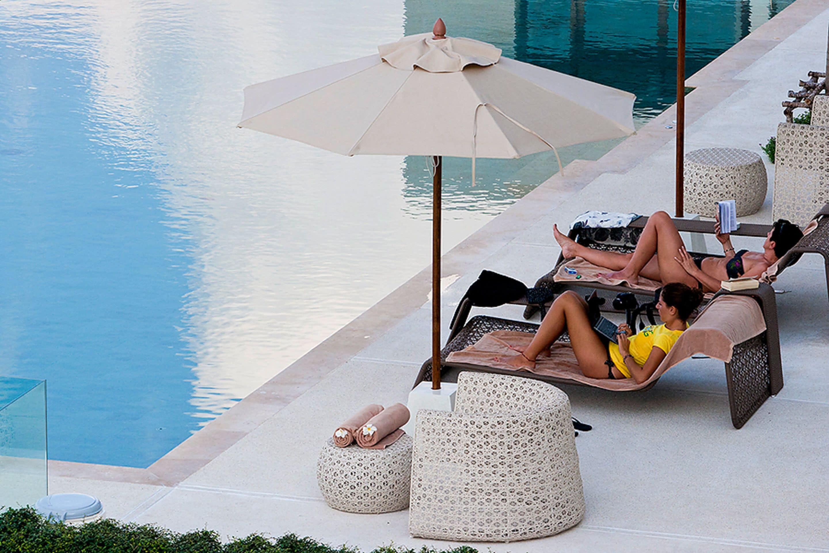 A poolside lounge area with a large umbrella, lounge chairs, and ottomans.