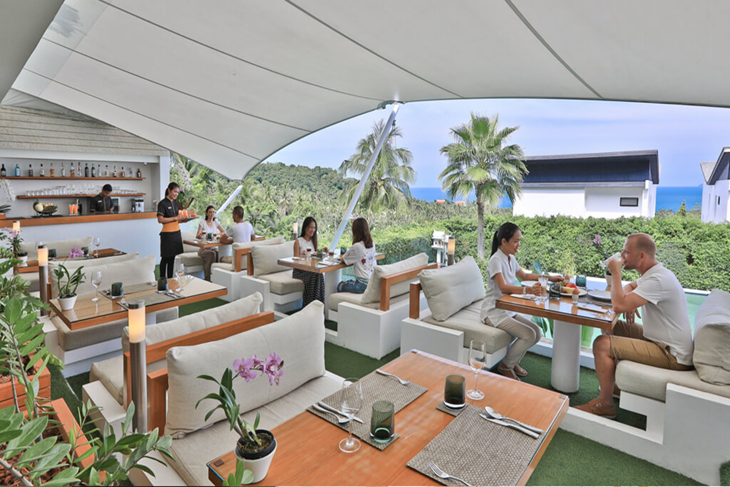 An outdoor dining area with multiple tables and chairs, a bar area with shelves of bottles, and a view of the ocean and hills in the background.