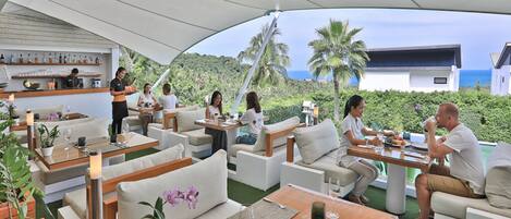 An outdoor dining area with multiple tables and chairs, a bar area with shelves of bottles, and a view of the ocean and hills in the background.