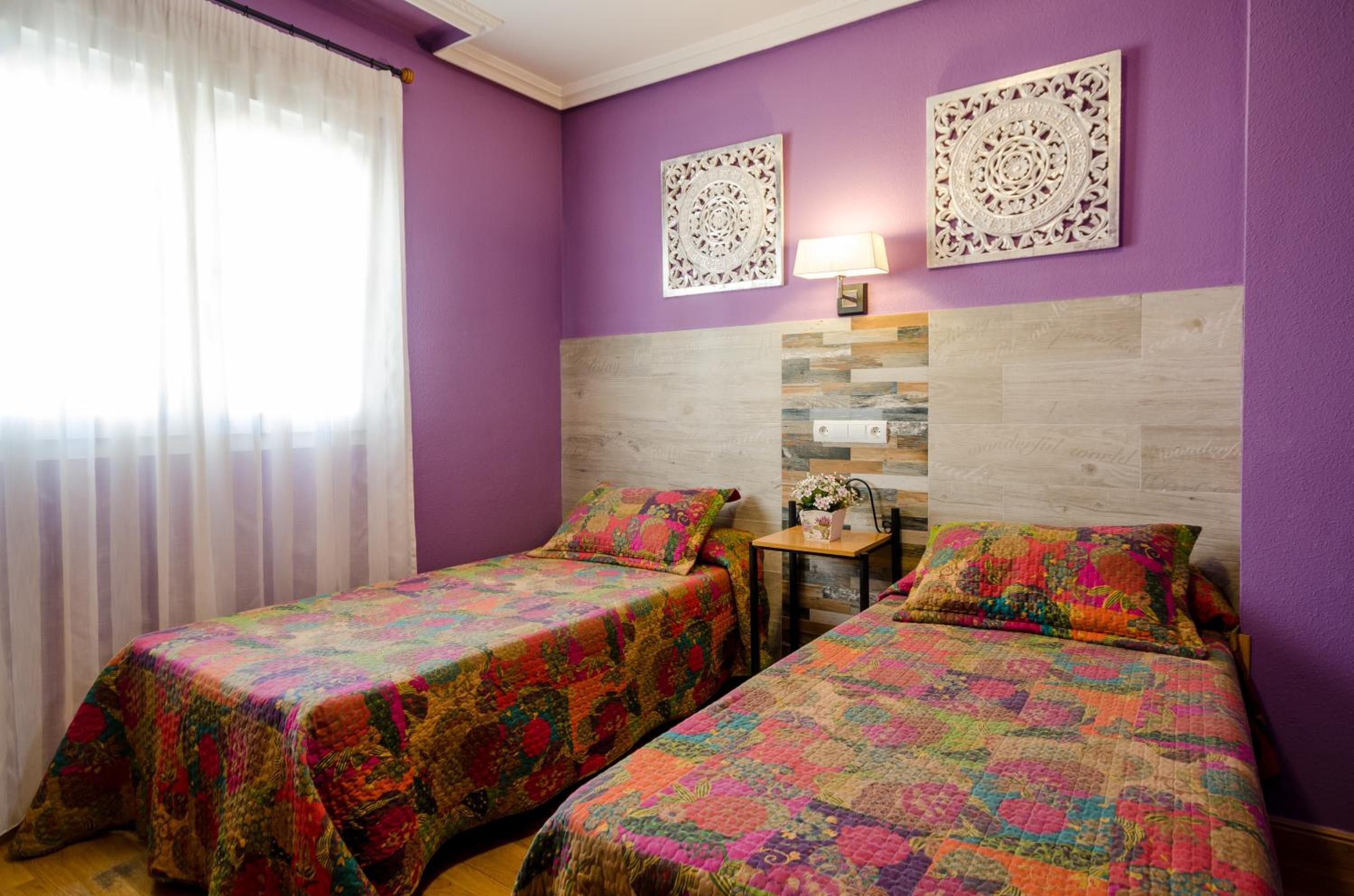 Quarto com duas camas, paredes roxas, detalhes decorativos nas paredes, uma mesinha com abajur e uma janela com cortinas leves.