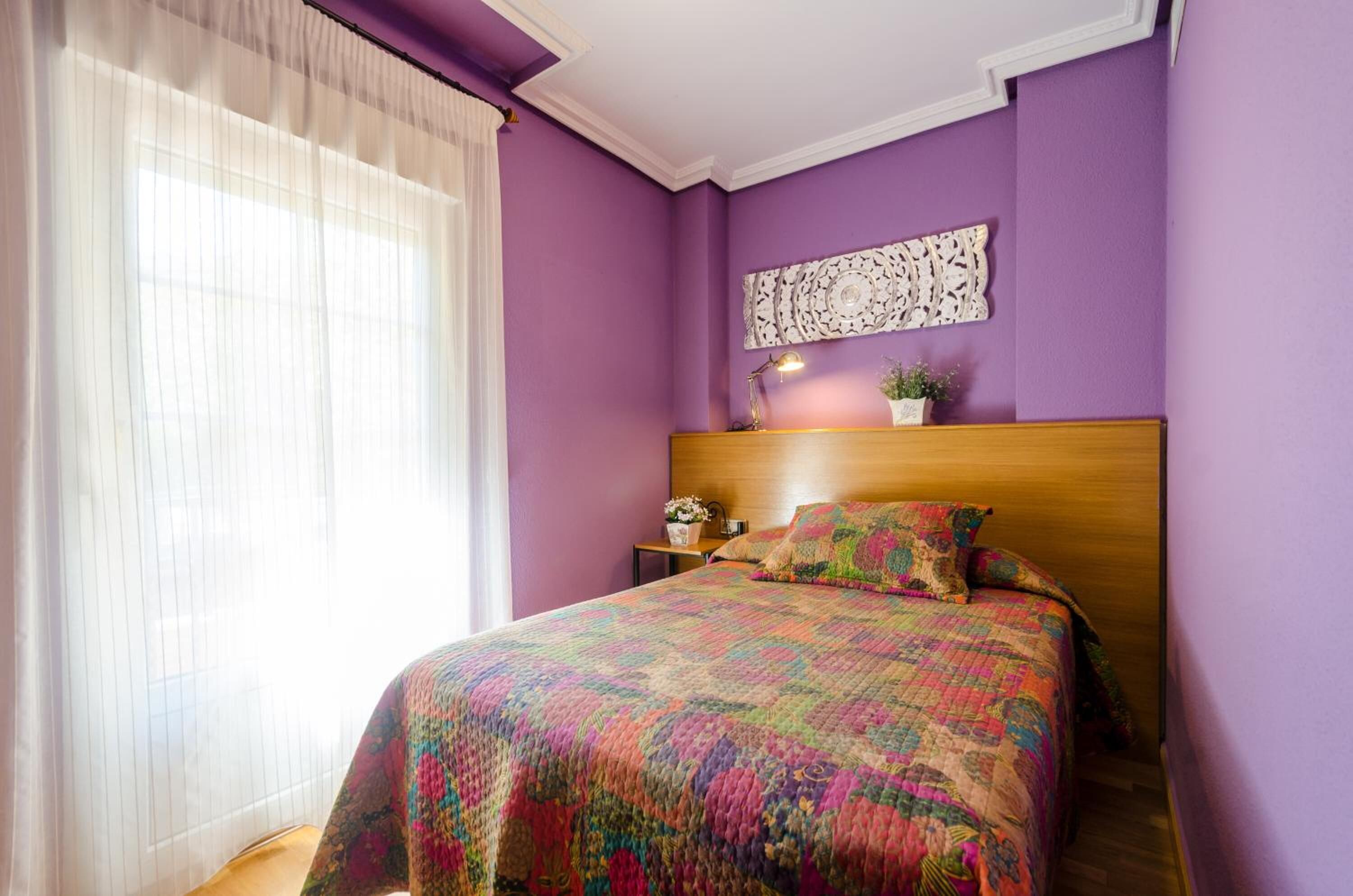 Un dormitorio con cabecera de madera, colcha colorida, mesita de noche con lámpara y un adorno en la pared.