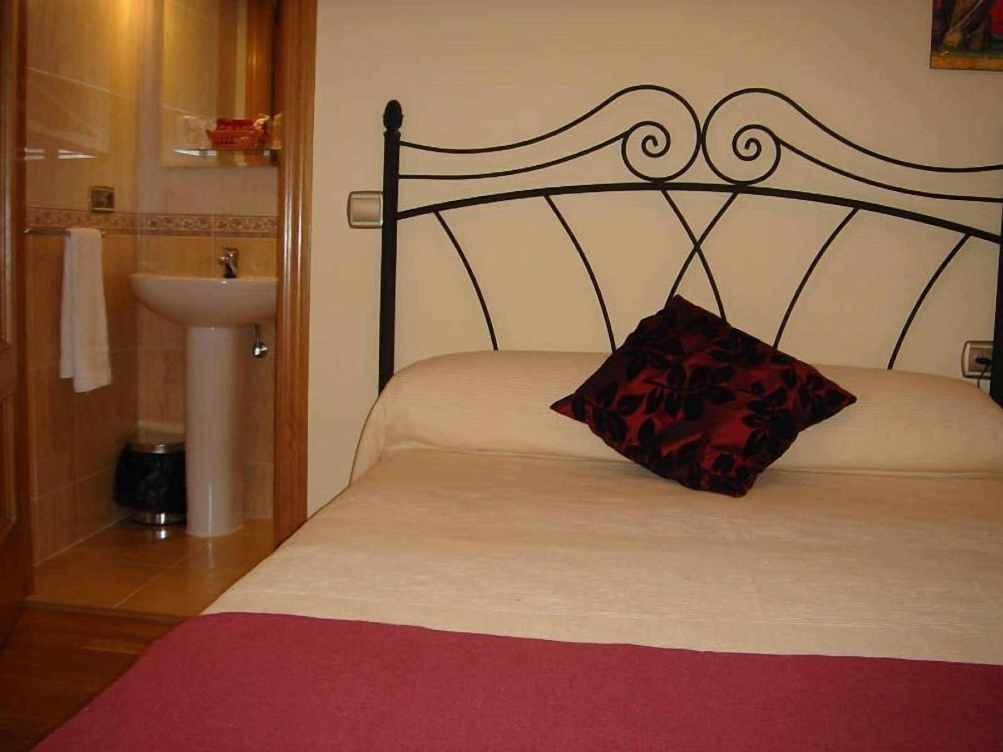 Un dormitorio con cama, almohada, mesita de noche, repisa en la pared y baño con lavamanos e inodoro.