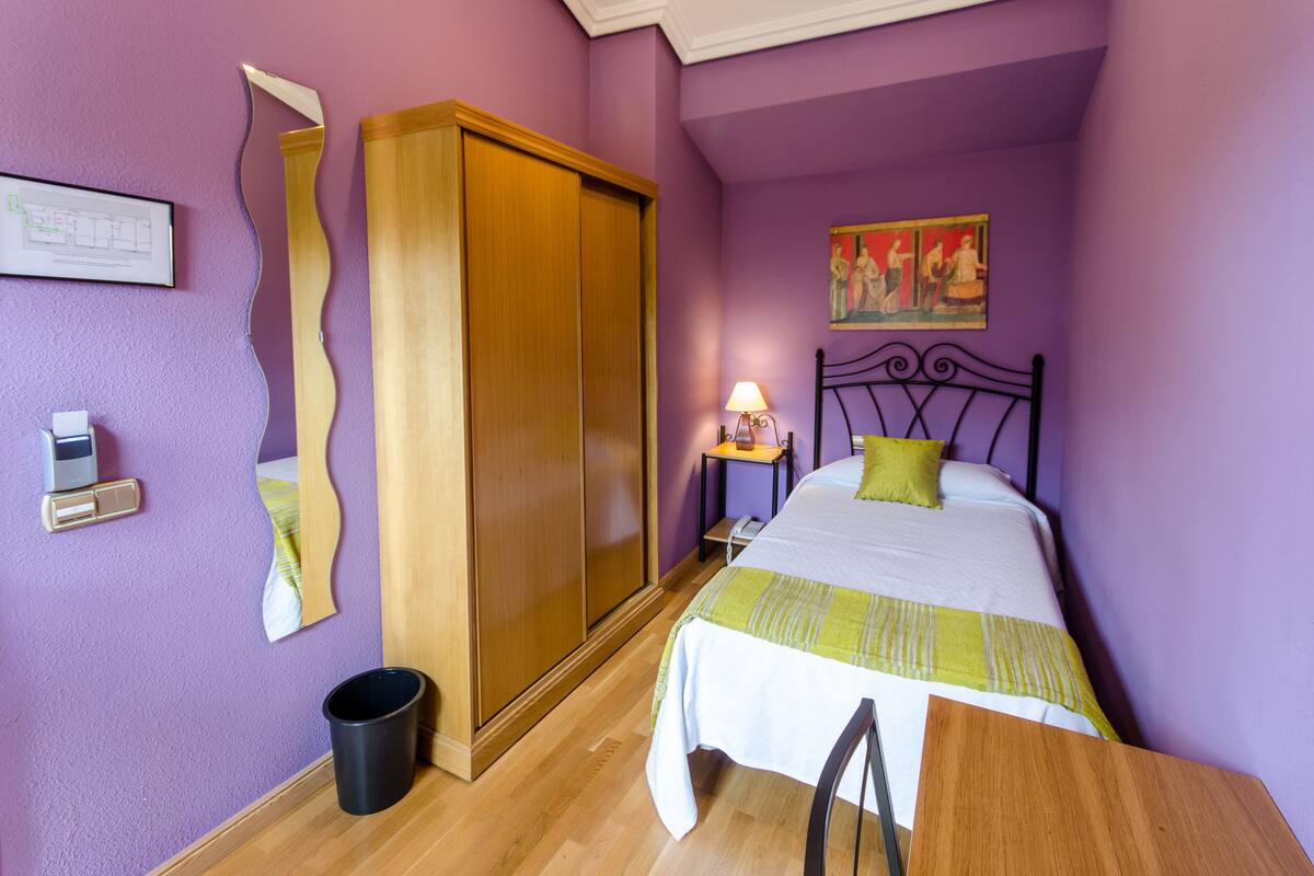 A bedroom with a bed, a wooden wardrobe, a bedside table with a lamp, and a painting on the wall.