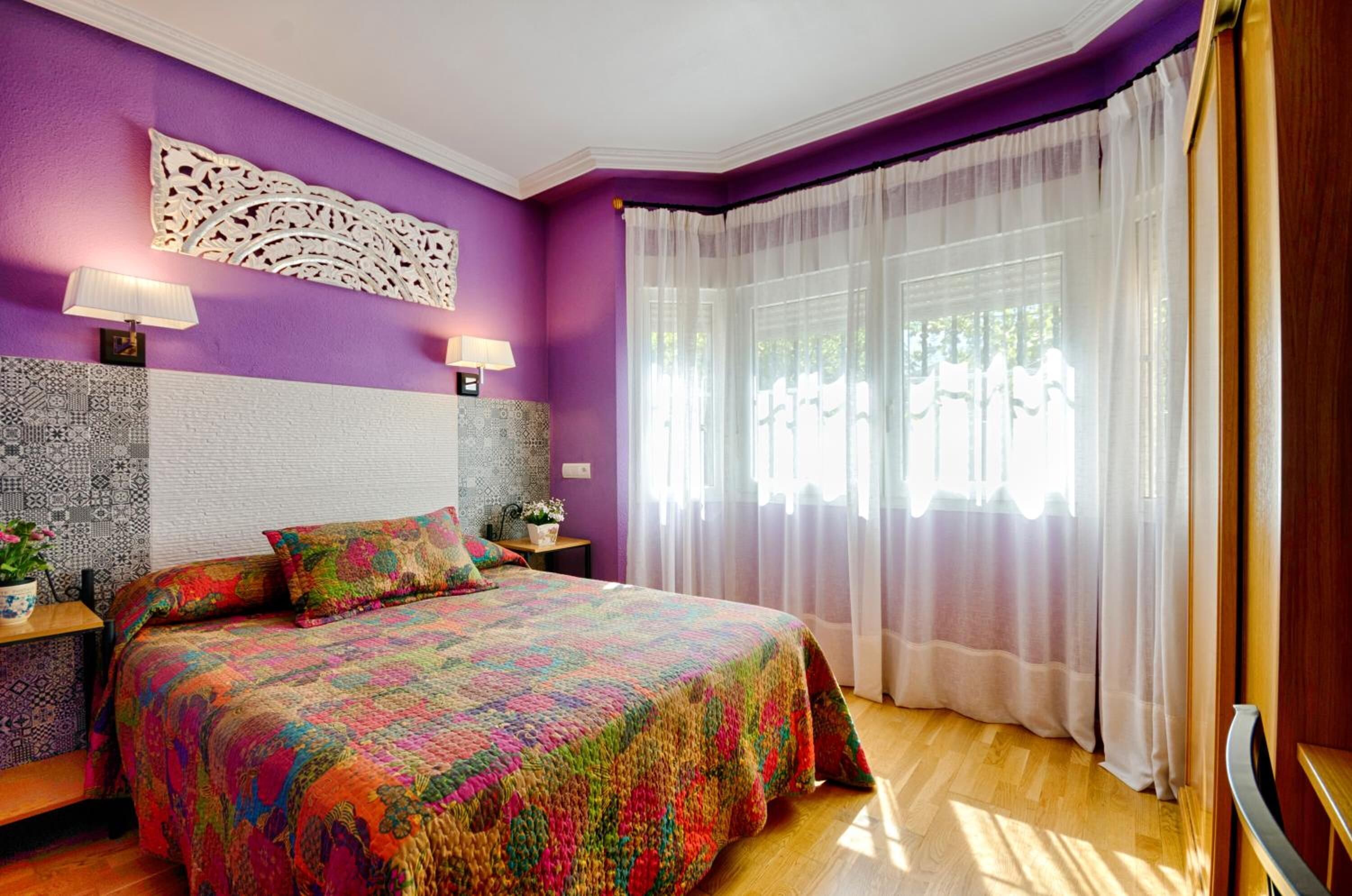 Une chambre à coucher avec une couvre-lit colorée à motifs matelassés, un panneau décoratif sur le mur et une grande fenêtre ornée de rideaux légers.