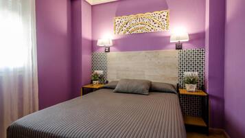 Quarto com cama, duas mesas de cabeceira, um objeto decorativo na parede e uma janela com cortinas translúcidas.
