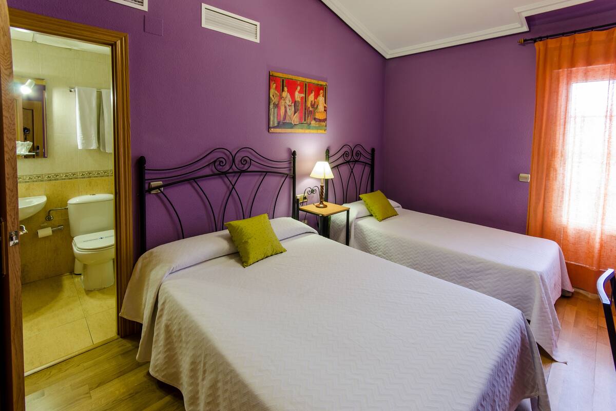 A room with two beds, a bathroom, and a painting on the wall.