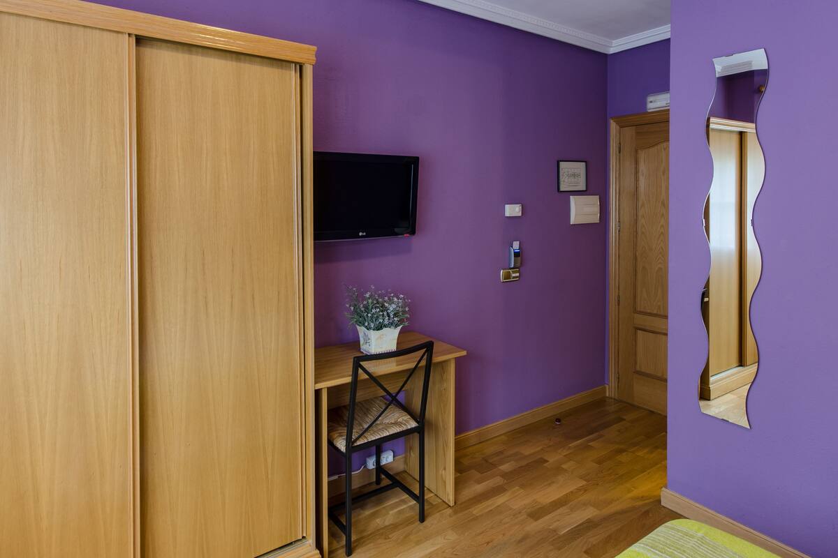 A room with a wooden desk, a chair, a television mounted on the wall, and a mirror on the wall.