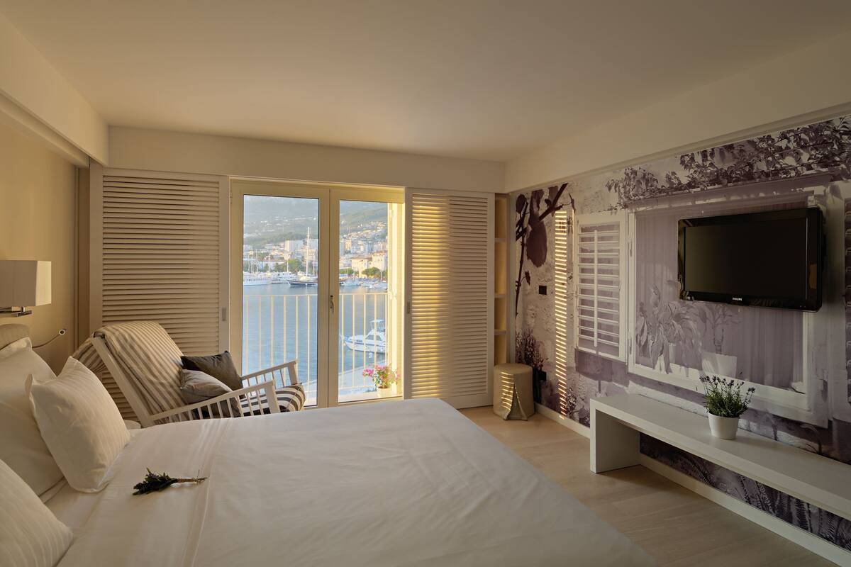 A hotel room with a large bed, a TV mounted on the wall, a balcony with a view of the city, and a potted plant on a console table.