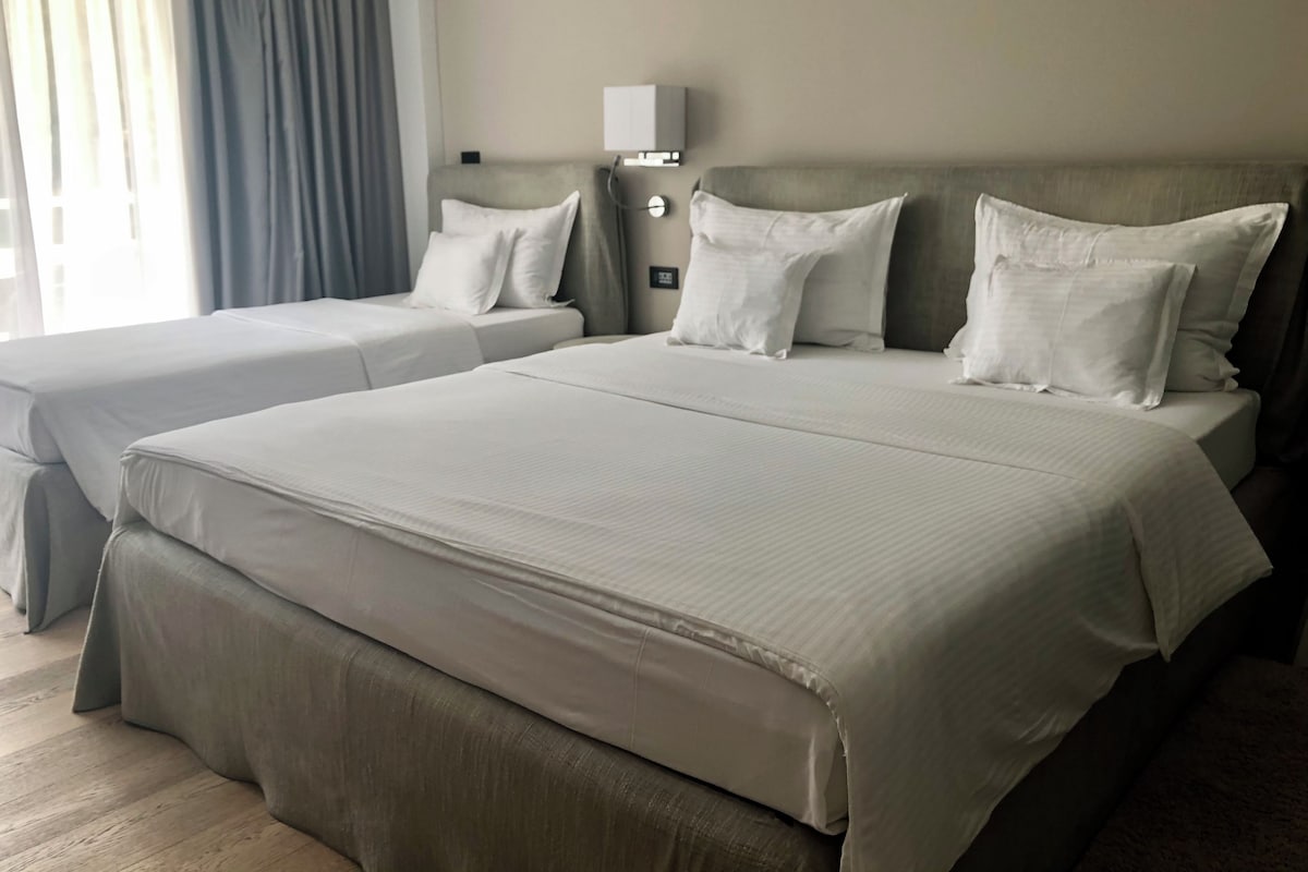 A hotel room with two beds, a bedside table with a lamp, and a window with curtains.