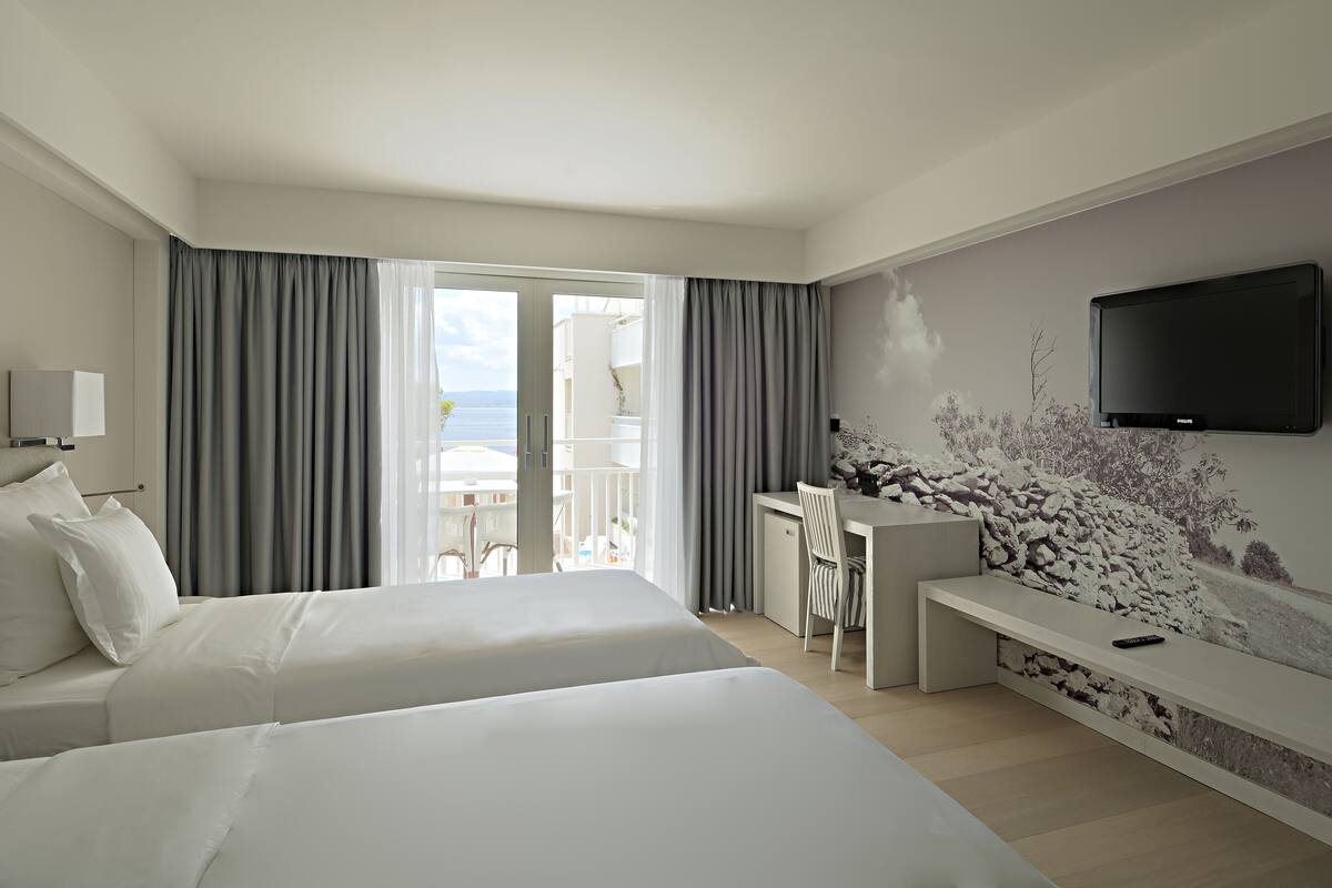A hotel room with two beds, a desk, a TV, and a balcony with a view of the sea.